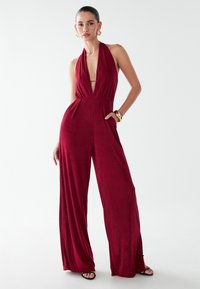 Mono burdeos con cuello halter, escote en V profundo, piernas anchas y bolsillos laterales. Tejido liso y texturizado con cintura ajustada. Sandalias negras.