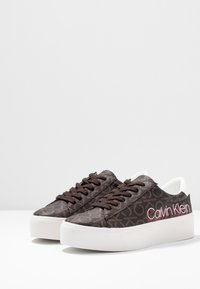 Sneakers Calvin Klein z brązowym wzorzystym wierzchem, białą podeszwą i ciemnymi sznurówkami. Posiadają logo w kolorze czerwonym oraz gładką teksturę. Niskoprofilowy design.
