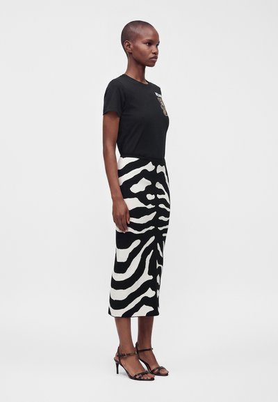 Femme debout de profil portant un t-shirt noir et une jupe midi motif zèbre noir et blanc avec des sandales noires à talons hauts.