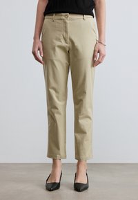 Pantalon beige droit porté avec des talons noirs à bout pointu et un haut noir, visible de la taille aux pieds sur un fond uni.
