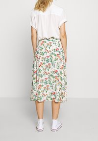 Jupe midi florale avec une base crème claire, ornée de fleurs vertes, rouges et bleues vives ; associée à des baskets blanches et une chemise à manches courtes.