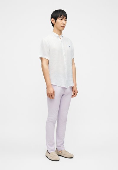 Άνδρας που στέκεται φορώντας ένα λευκό πουκάμισο με κοντά μανίκια και κουμπιά, ανοιχτόχρωμο λιλά παντελόνι και μπεζ μοκασίνια, με φόντο άσπρο και απλό.