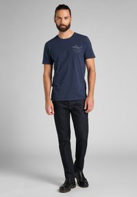 Camiseta de algodón azul marino con mangas cortas y escote redondo, que presenta un pequeño logo de "Genuine Jeans" en el lado izquierdo del pecho. Jeans oscuros y botas negras.