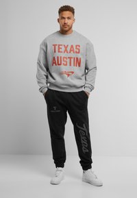 Sweatshirt gris avec "TEXAS AUSTIN" en lettres rouges en gras. Pantalons de survêtement noirs avec "Texas" imprimé le long de la jambe. Baskets blanches, coupe décontractée.