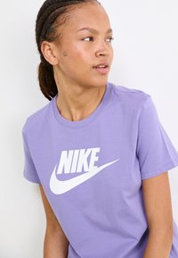 Vijolična bombažna majica s kratkimi rokavi, ki ima na prsih velik bel Nike logo, okroglo ovratnico in je prikazana na svetli podlagi.