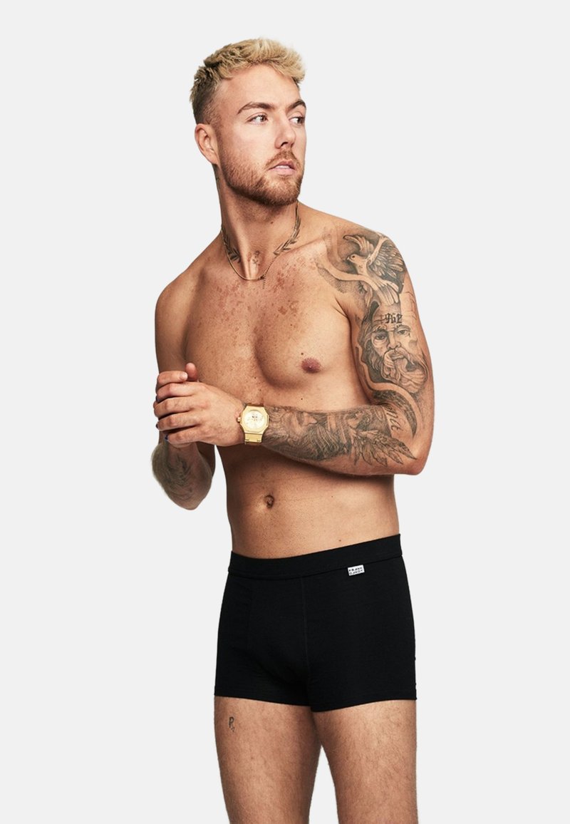 Svarta boxertrosor med en mjuk, elastisk midjeband; modellen har synliga tatueringar, kort ljust hår och en guldfärgad klocka på handleden.