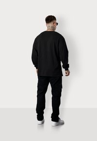 Schwarzes Langarmshirt und Cargo-Hose, beide in Schwarz. Das Shirt hat eine lockere Passform; die Hose verfügt über Seitentaschen. Weiße Sneakers vervollständigen das Outfit.