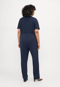 Navy jumpsuit with a relaxed fit, short sleeves, elastic waistband, and wide legs. Smooth fabric with a matte texture, paired with patterned heels.