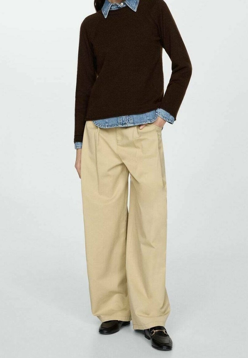 Personne portant un pantalon beige à jambes larges, des mocassins noirs, une chemise en jean bleue et un pull marron foncé, debout avec une main dans la poche.