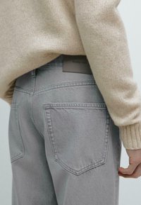 Gray denim jeans with two back pockets, a leather label on the waistband, paired with a beige knit sweater. Textured knit and relaxed fit.
