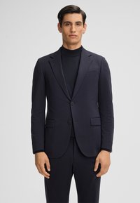 Blazer blu navy con un motivo testurizzato, design a un petto, revers a punta e due tasche frontali, indossato sopra un dolcevita abbinato.