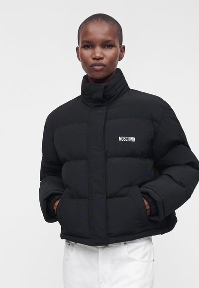 Personne portant une doudoune noire Moschino avec les mains dans les poches, associée à un pantalon blanc, sur un fond uni.