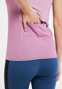 Svijetloroza sportska majica s teksturiranom površinom i crnom zatvaračnom džepom na leđima. Uparena s tamnoplavim tajicama.