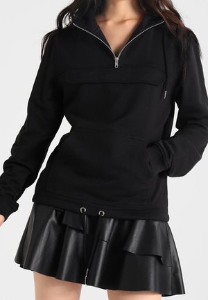 Sort sweatshirt med halv lynlås, frontlomme og hætte. Kombinationen inkluderer et sort nederdel i kunstlæder med flæse lag og snorejusteringer.