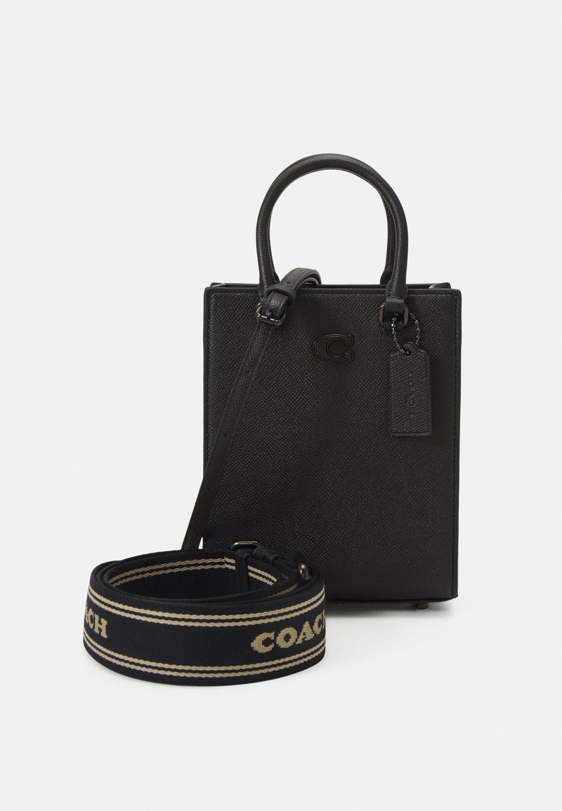 Sac à main Coach en cuir noir avec poignées supérieures, une large bandoulière en tissu amovible avec le texte "Coach" et une petite étiquette en cuir suspendue.