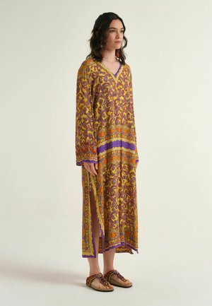 Donna in piedi che indossa un lungo vestito giallo ampio con motivi paisley viola e arancioni e sandali abbinati su uno sfondo semplice.
