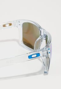 Ochelari de soare transparenți cu lentile degrade maro, accente albastre pe tâmple și un design elegant, modern, având un cadru semi-fără ramă.
