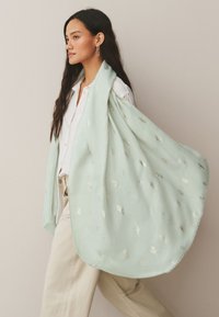 Mujer con pelo largo y oscuro que lleva pendientes de oro, camisa blanca, pantalones beige y una bufanda azul claro con un sutil patrón de hojas.
