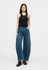 Νεαρή γυναίκα στέκεται με το ένα χέρι στην τσέπη, φορώντας μαύρο αμάνικο τοπ, φαρδιά μπλε τζιν παντελόνια και σκούρα καφέ chunky μποτάκια.