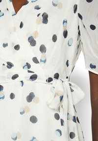 Robe portefeuille en tissu léger et texturé avec une base blanche et des motifs circulaires variés en bleu, noir et beige. Se noue à la taille.