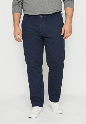 Mand iført marineblå straight-fit bukser, grå langærmet skjorte og hvide sneakers, stående mod en hvid baggrund.