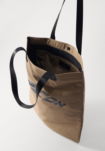 Brown fabric tote bag with a textured surface, featuring a black logo, long black straps, and an open top design. Interior is lined in black.