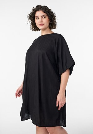 Žena s kučeravými vlasmi, oblečená v voľných čiernych šatách po kolená s krátkymi nadýchanými rukávmi, stojaca na jednoduchom bielom pozadí.