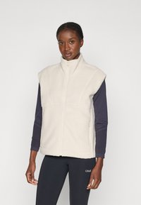 Fleece vest in crèmewit met een hoge kraag, ritssluiting en schoudernaden. Draagt over een marineblauw shirt met lange mouwen. Vlakke stofstructuur.