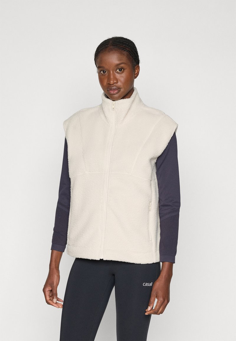 Fleece vest in crèmewit met een hoge kraag, ritssluiting en schoudernaden. Draagt over een marineblauw shirt met lange mouwen. Vlakke stofstructuur.