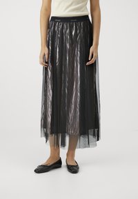 Gonna di tulle nera con finitura metallizzata, caratterizzata da una ampia vita elastica, design fluido e texture a strati. Indossata con ballerine nere.