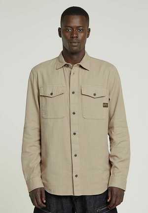 Beige langslavenshirt met twee borstzakken, knoopsluiting en een stoffen label. Gelaagd materiaal en eenvoudig ontwerp met donkere knopen.