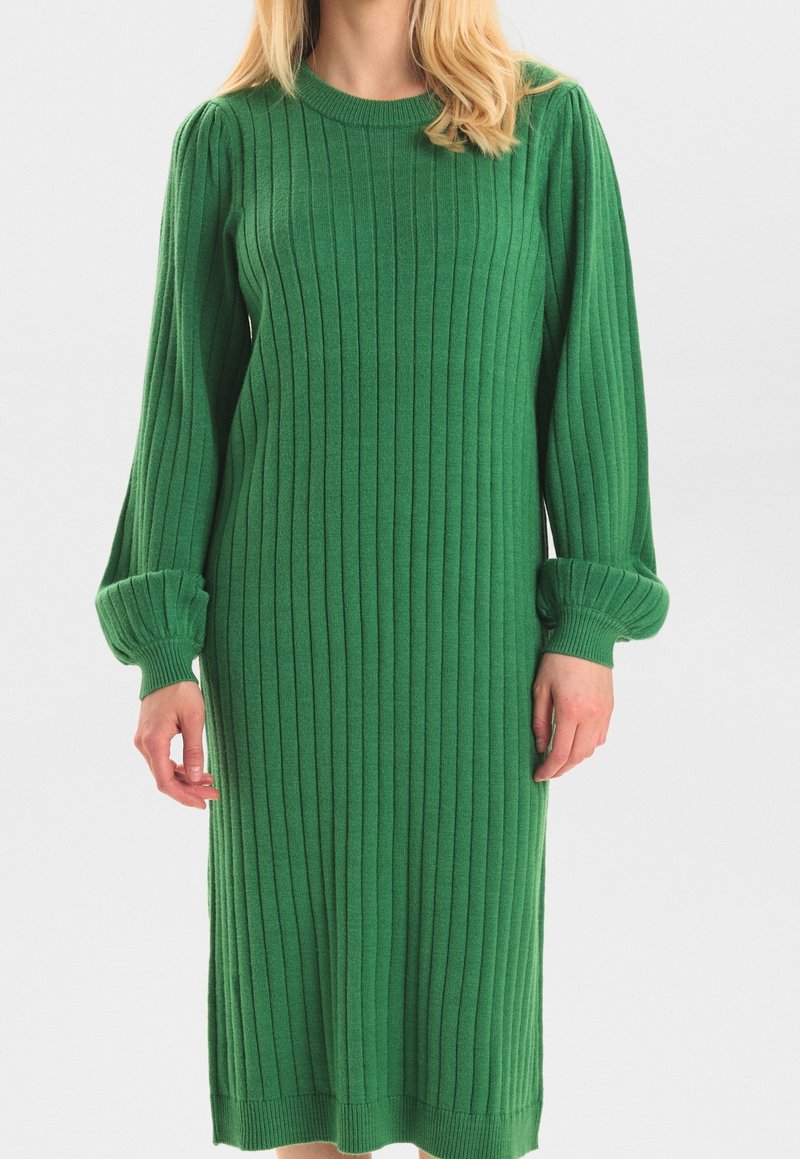 Femme portant une longue robe en maille côtelée verte avec des manches longues bouffantes et des poignets ajustés, sur un fond clair uni.