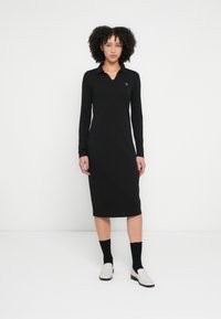 Schwarzes langärmeliges Kleid mit Polokragen, figurbetonter Silhouette, knielang, glatter Textur, mit kleinem Logo auf der linken Brust.