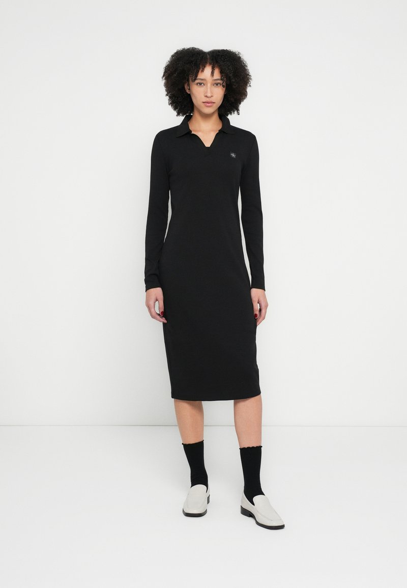 Schwarzes langärmeliges Kleid mit Polokragen, figurbetonter Silhouette, knielang, glatter Textur, mit kleinem Logo auf der linken Brust.