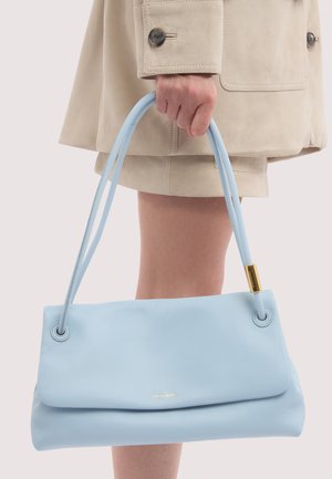 Hand hält eine hellblaue Ledertasche mit doppelten Griffen neben einem beigefarbenen Mantel und Rock.