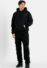 Czarna bluza z kapturem i kieszenią kangurką, w połączeniu z czarnymi spodniami cargo z wieloma kieszeniami i zamkami błyskawicznymi. Uzupełnione czarnymi sneakersami.