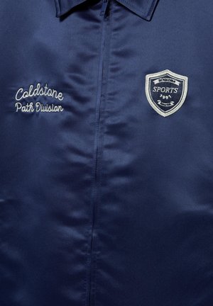 Mørkeblå jakke med lynlås foran, broderet tekst "Coldstone Path Division" på venstre side, og sportsemblem på højre bryst.