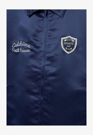 Granatowa kurtka z zamkiem z przodu, z nadrukiem "Coldstone Path Division" po lewej stronie oraz naszywką emblematową sekcji sportowej na prawym ramieniu.