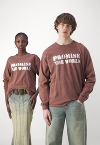 Brun langermet skjorte med "LOVER VERDEN" i hvit tekst, kombinert med et grønn-blått denimskjørt og avslappede jeans.