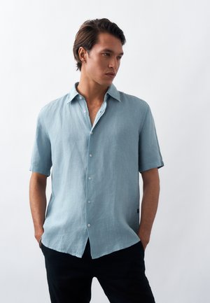 Νέος άνδρας με κοντά σκούρα μαλλιά, φορώντας ένα ανοιχτό γαλάζιο κοντομάνικο πουκάμισο με κουμπιά και μαύρο παντελόνι, στέκεται με τα χέρια στις τσέπες.