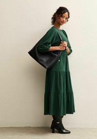 Kobieta w długiej zielonej sukience i czarnych butach na obcasie, trzymająca dużą czarną torbę na ramię, stoi przy beżowej ścianie.