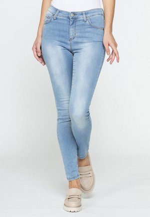 Donna che indossa jeans skinny azzurri chiari e scarpe slip-on beige, in piedi contro uno sfondo bianco semplice.