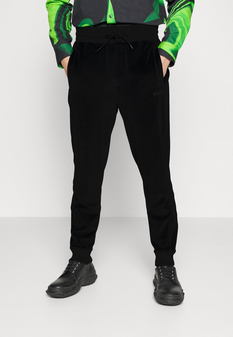 Homme portant un pantalon de jogging noir et des chaussures à lacets noires, les mains dans les poches, avec une chemise boutonnée à motifs verts.