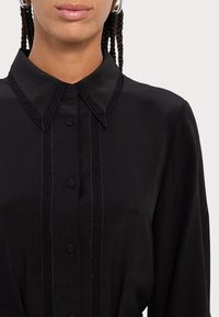 Melna blūze ar apkakles dekoltē, ar mežģīņu apdari gar pogu slīdi un piedurknēm, mīksts audums un pieguļoša silueta.