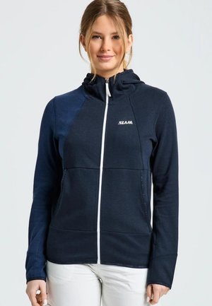 Una mujer luce una chaqueta azul marino con capucha, cremallera blanca en la parte delantera y logotipo, combinada con unos pantalones blancos sobre un fondo claro.