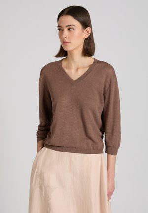 Vrouw met recht bruin haar, een bruine v-hals trui en een lichtbeige rok met borduursel, handen in de zakken, kijkt naar de zijkant.