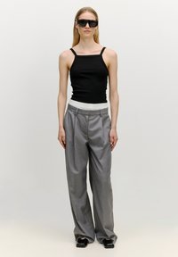 Schwarzes geripptes Tanktop mit dünnen Trägern, hochgeschnittene graue Plisseehosen und klobige schwarze Schuhe. Das Model trägt zudem übergroße schwarze Sonnenbrillen.