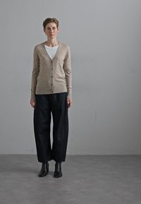 Beige Strickjacke mit Knöpfen, getragen über einem weißen Hemd. Dunkle, lockere Hose und schwarze Stiefeletten runden das Outfit ab. Glatte Textur.