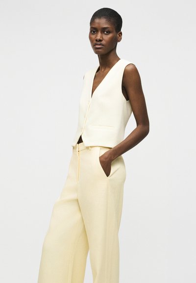 Γυναίκα που στέκεται με το ένα χέρι στην τσέπη, φορώντας αμάνικο εκρού γιλέκο και ταιριαστό ψηλόμεσο παντελόνι φαρδιάς γραμμής, μπροστά σε απλό φόντο.