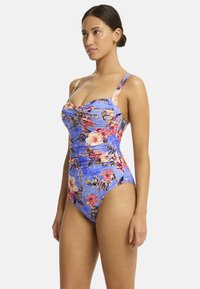Model poseert in een bloemen badpak met een blauwe achtergrond, met een ruchesontwerp en brede schouderbanden.